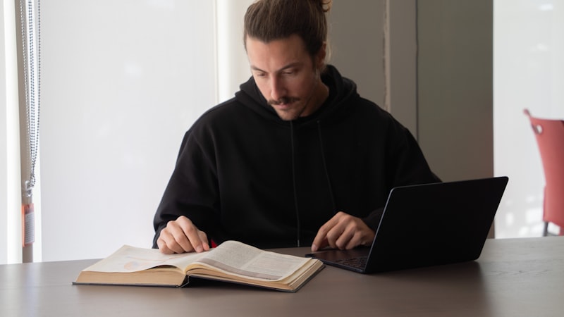 A man reading a book while using his laptop to learn a new skill