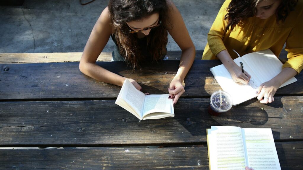 Two people reading books and writing in a notebook at a picnic table