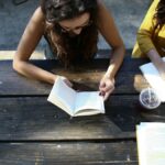 Two people reading books and writing in a notebook at a picnic table